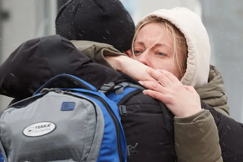 A woman with a hood hugs another person tightly, her eyes filled with emotion. Snow falls around them, and the person she’s hugging wears a backpack and a black beanie.