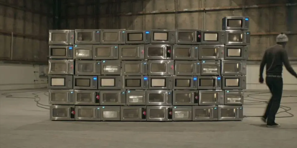A person in a beanie walks past a large stacked wall made of multiple stainless steel microwave ovens inside a spacious, industrial-style room.