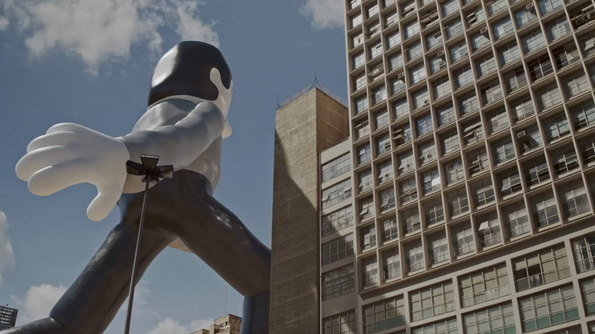 A giant cartoon-like statue of a black-and-white figure walks between tall city buildings under a blue sky with scattered clouds.