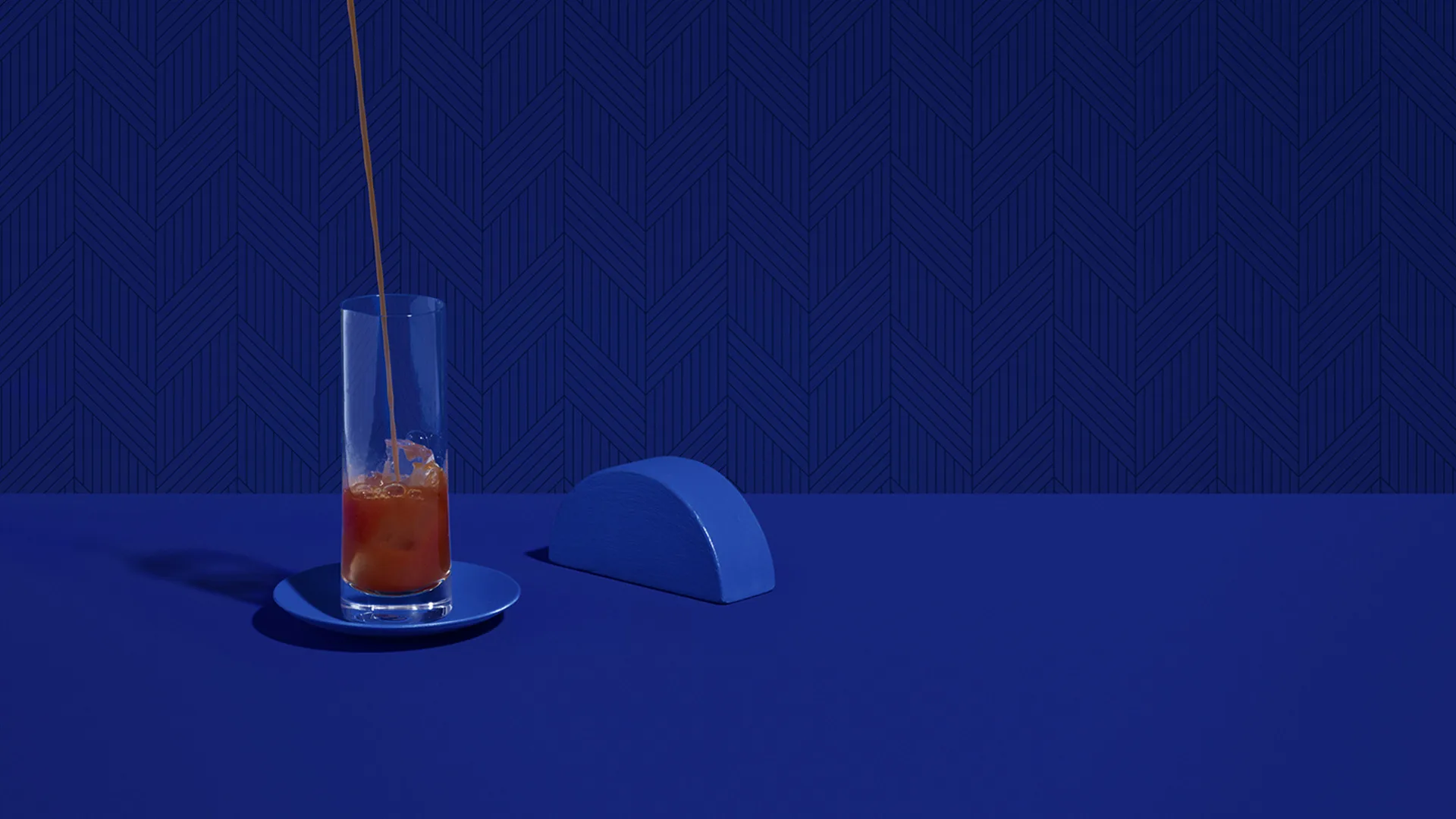A stream of orange-brown liquid is being poured into a tall glass on a blue saucer, set on a blue surface with a geometric blue background and a blue semicircular object beside the glass.