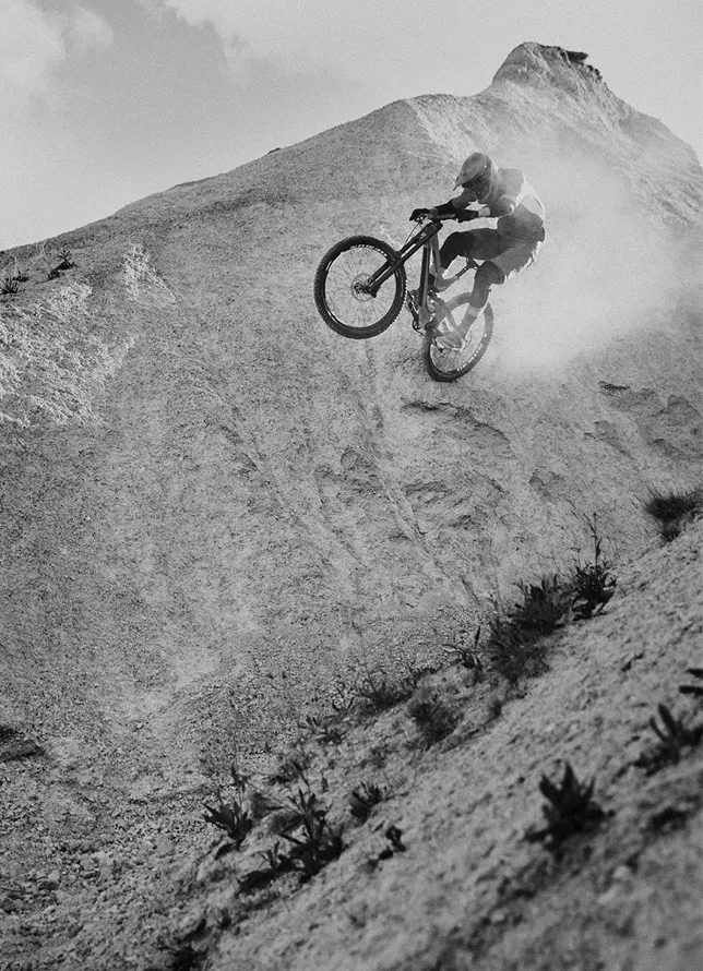 A mountain biker in mid-air performs a steep descent down a rocky slope, kicking up dust. The scene is captured in black and white, emphasizing the dramatic landscape and motion.