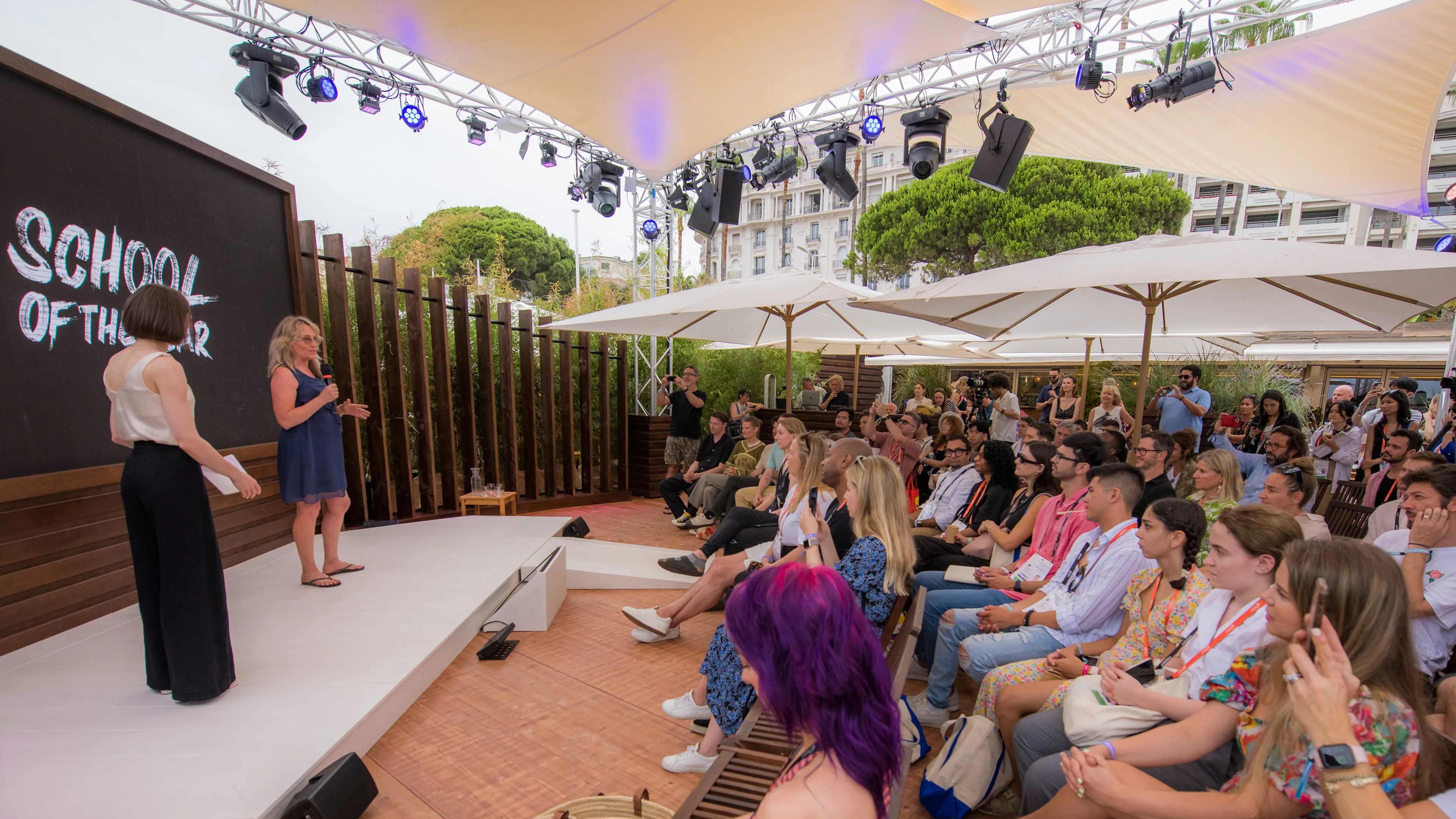 A woman speaks on an outdoor stage labeled School of the Unknown to a seated audience under large umbrellas, with event lighting and buildings visible in the background.