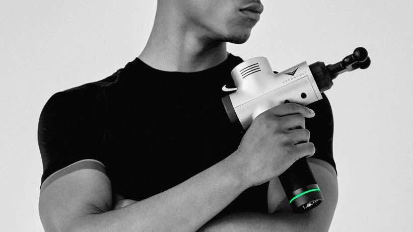 A person in a black t-shirt uses a handheld massage gun on their shoulder, with the device held across their chest. The image is in grayscale, and the persons face is partially out of frame.