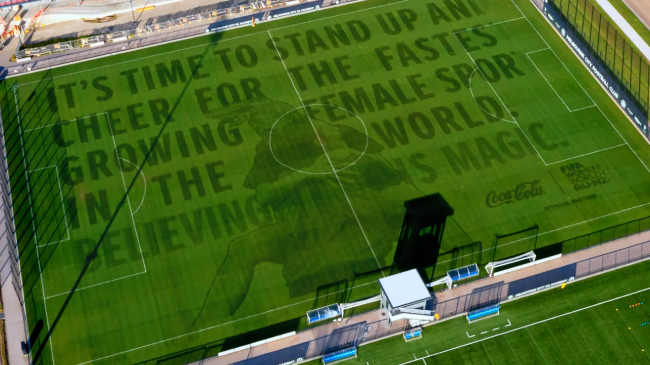 Aerial view of a soccer field with text on the grass that reads: ITS TIME TO STAND UP AND CHEER FOR THE FASTEST GROWING FEMALE SPORT IN THE WORLD. BELIEVE IN ITS MAGIC. Logos for Coca-Cola and FIFA Womens World Cup are visible.