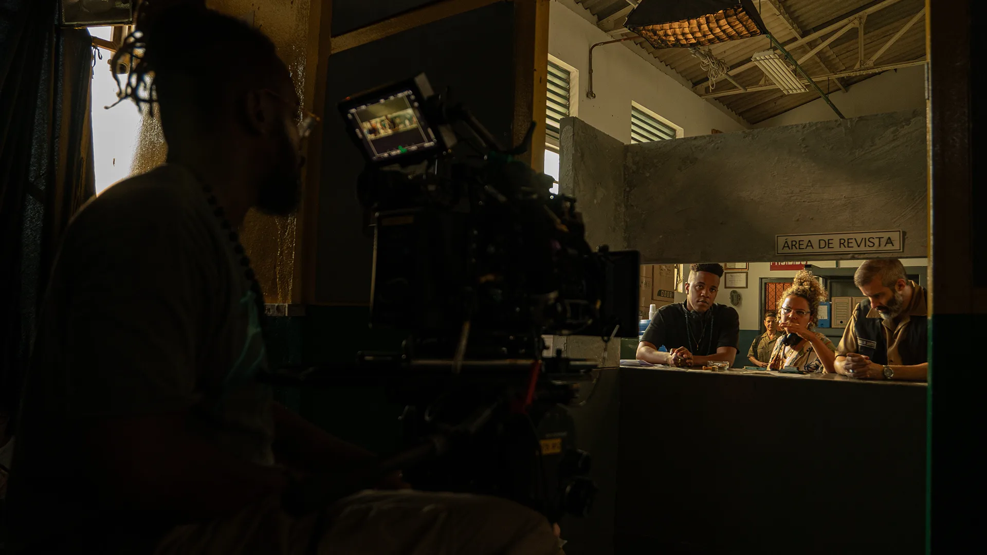 A camera operator films four people talking at a counter in a dimly lit room marked Área de Revista. The set has industrial decor with visible ceiling beams and windows.