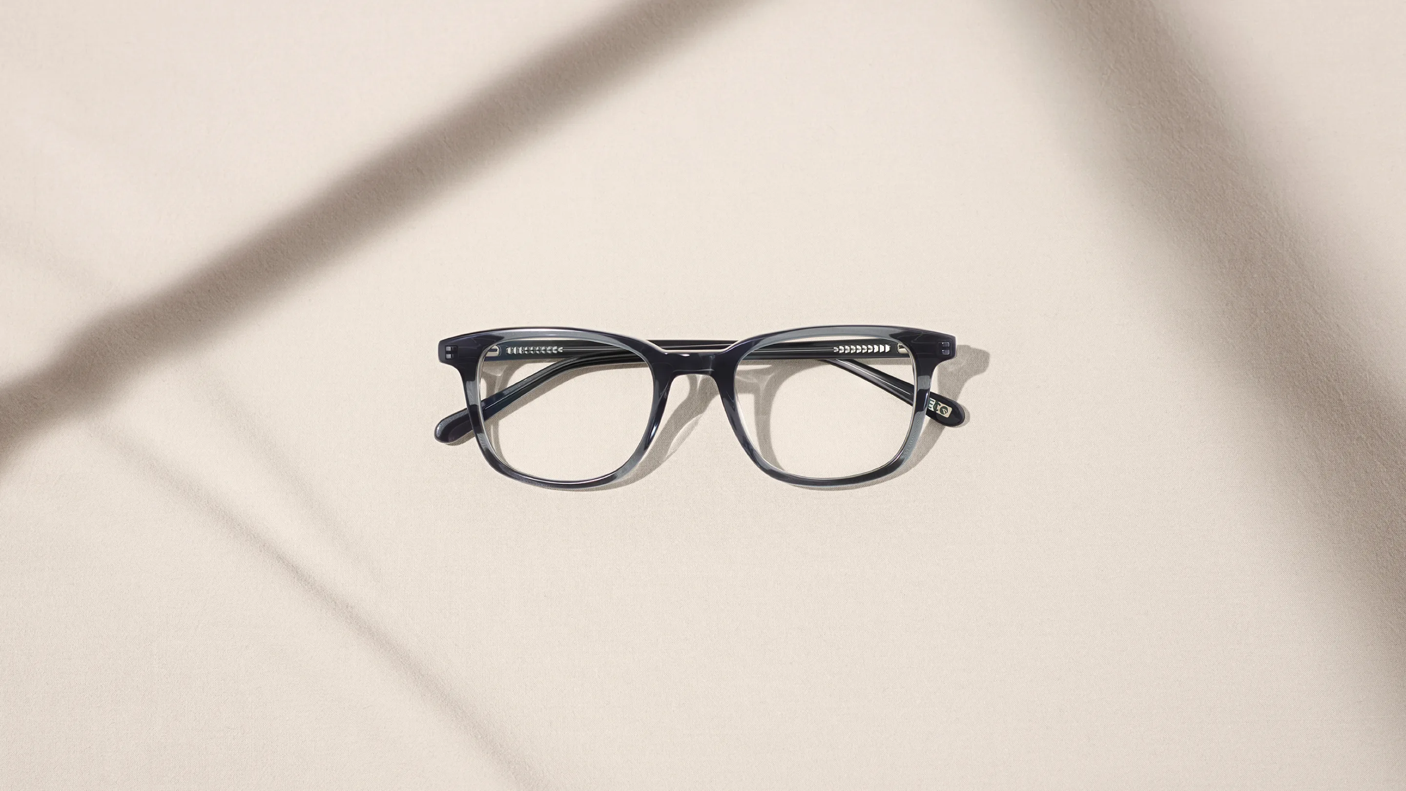 A pair of black-rimmed eyeglasses is centered on a light beige surface with soft shadows crossing diagonally in the background.