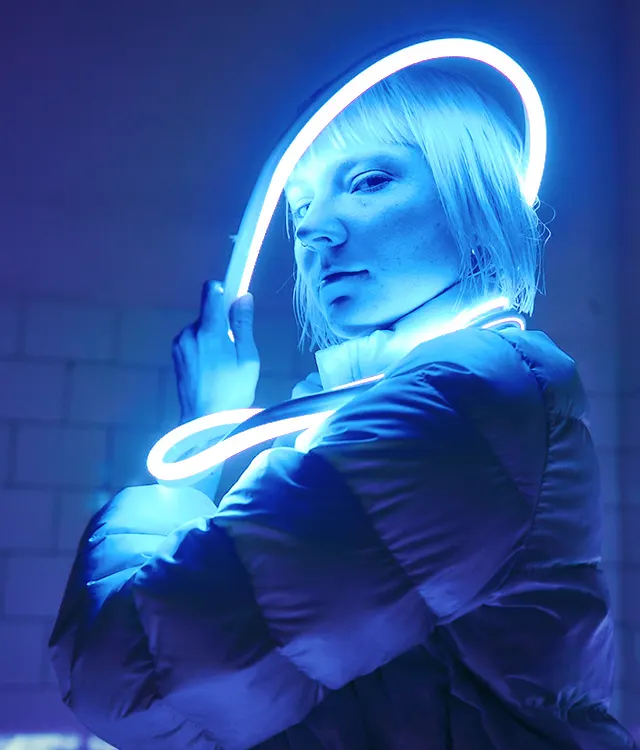 A person with short, light-colored hair is wrapped in a glowing blue neon light, wearing a puffy jacket, and standing against a tiled wall in a dimly lit setting.