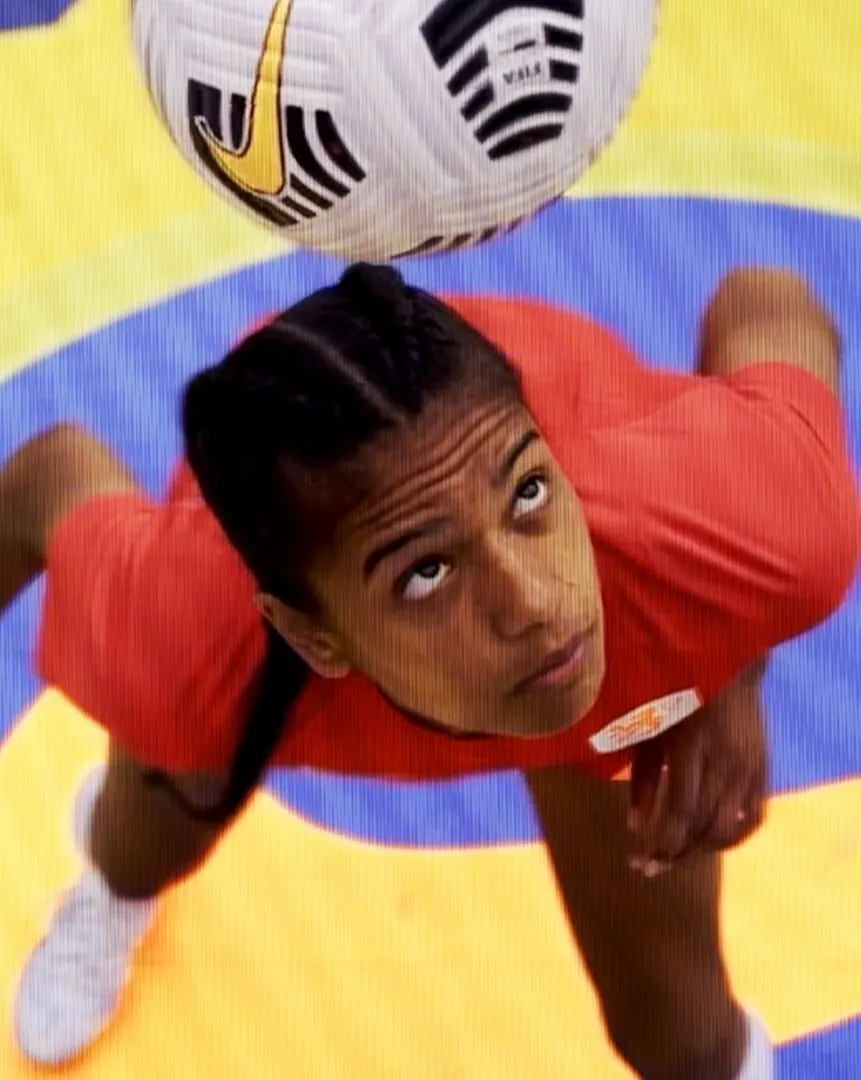 A soccer player in a red shirt and white shoes balances a black-and-white soccer ball on their head, looking up with focus on a yellow and blue court.