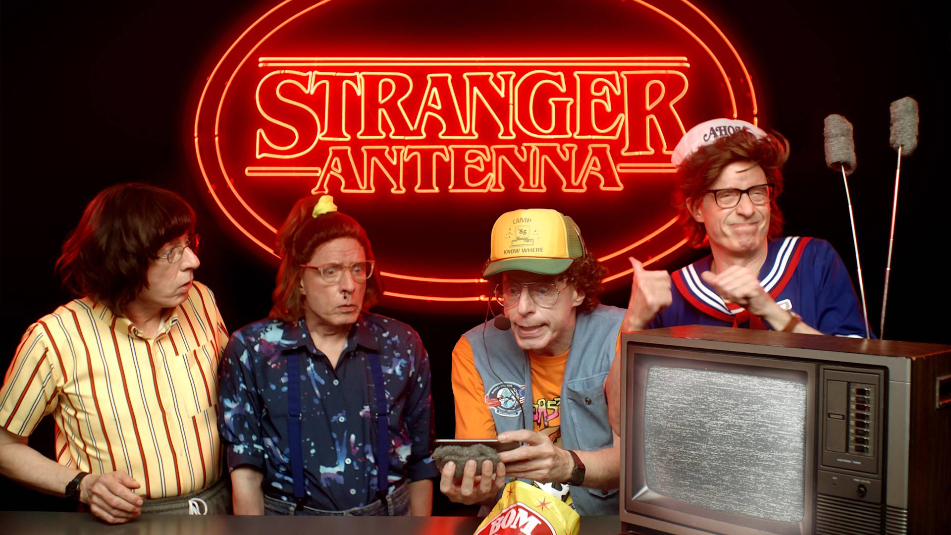Four people dressed in retro outfits stand in front of a neon sign reading “Stranger Antenna.” One holds a remote, another wears a sailor hat, and an old TV with static is in the foreground. The scene is quirky and comedic.