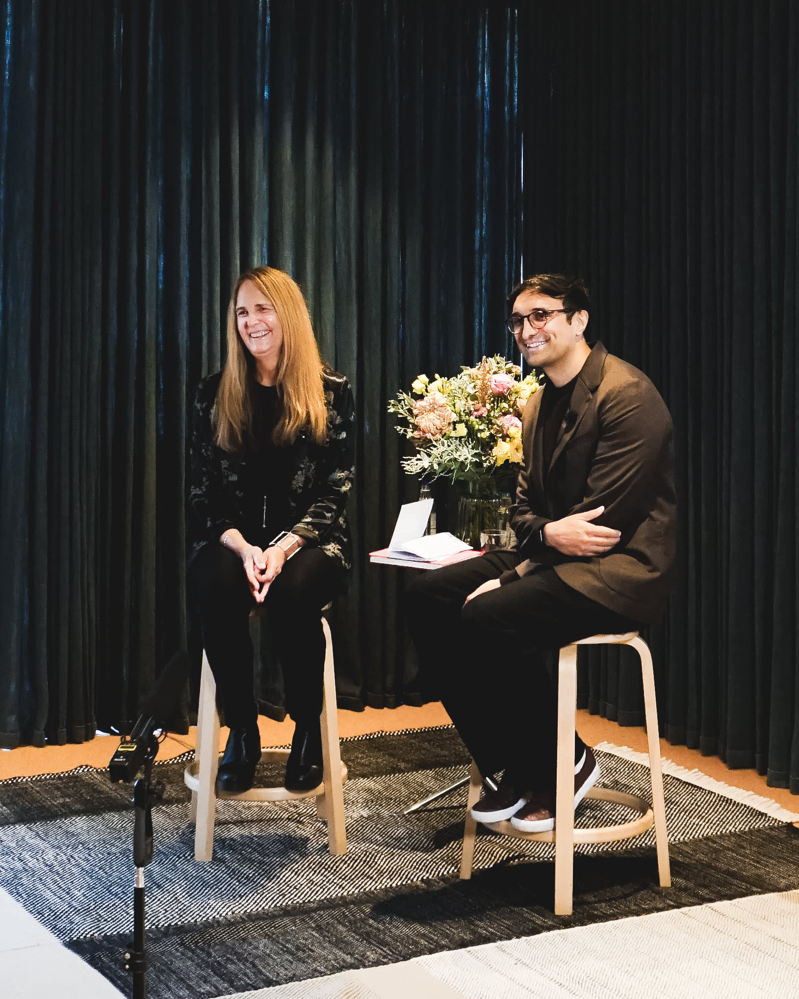Two people sit on stools in front of dark curtains. The woman on the left is smiling and the man on the right is holding papers, also smiling. A bouquet of flowers is on a table between them. A microphone is visible in the foreground.