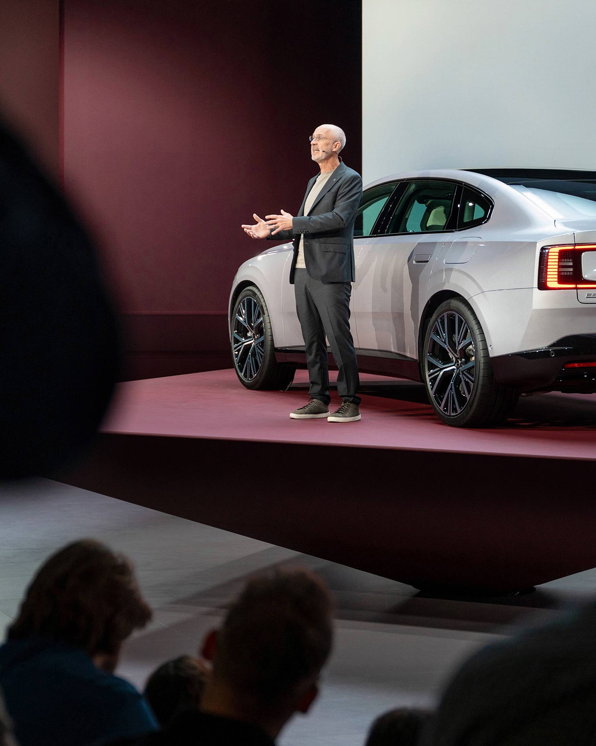 A man in a suit stands on a stage next to a silver car, speaking to an audience at a presentation or car launch event in a modern, well-lit venue.