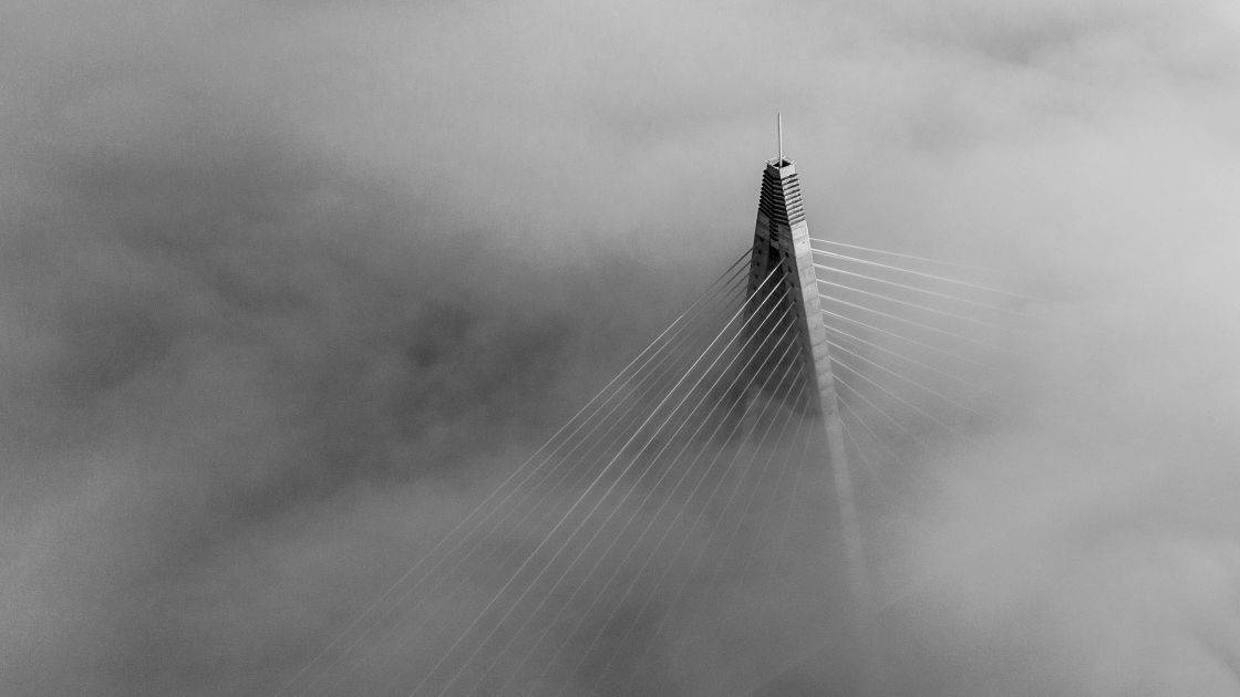 A cable-stayed bridge tower rises through dense fog, with suspension cables fanning out and disappearing into the mist, creating a mysterious and dramatic atmosphere in black and white.