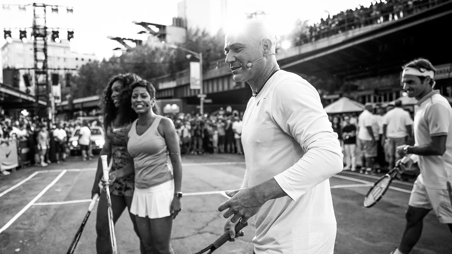 Three tennis players on an outdoor court smile and interact as a crowd watches in the background. The photo is in black and white, creating a lively, energetic scene at a street tennis event.