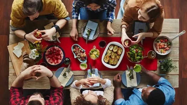 A group of six people sit around a table sharing a meal, dishes including salads, vegetables, and drinks. The scene is viewed from above, with a red table runner and a friendly, communal atmosphere.