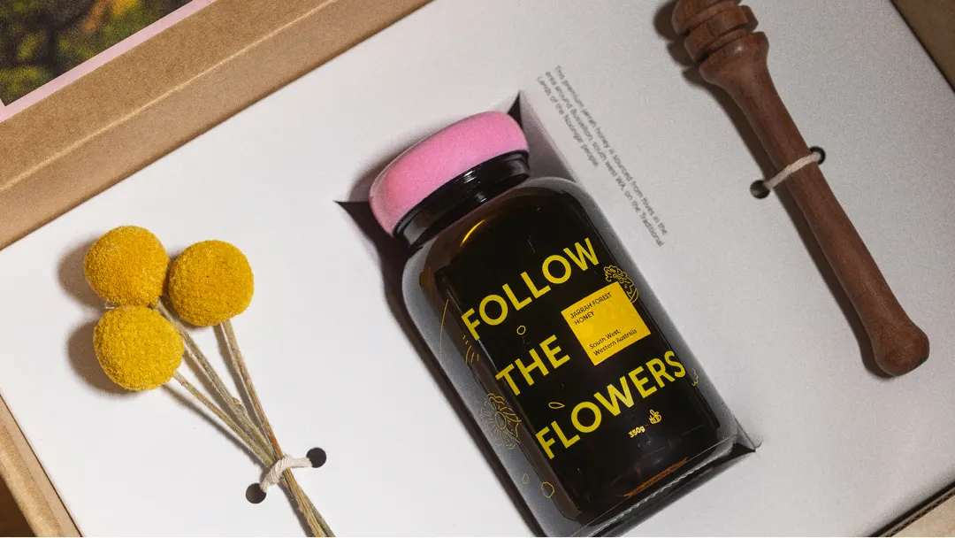 A gift box with yellow billy ball flowers, a glass jar labeled Follow the Flowers with a pink lid, and a wooden honey dipper placed on a white insert.