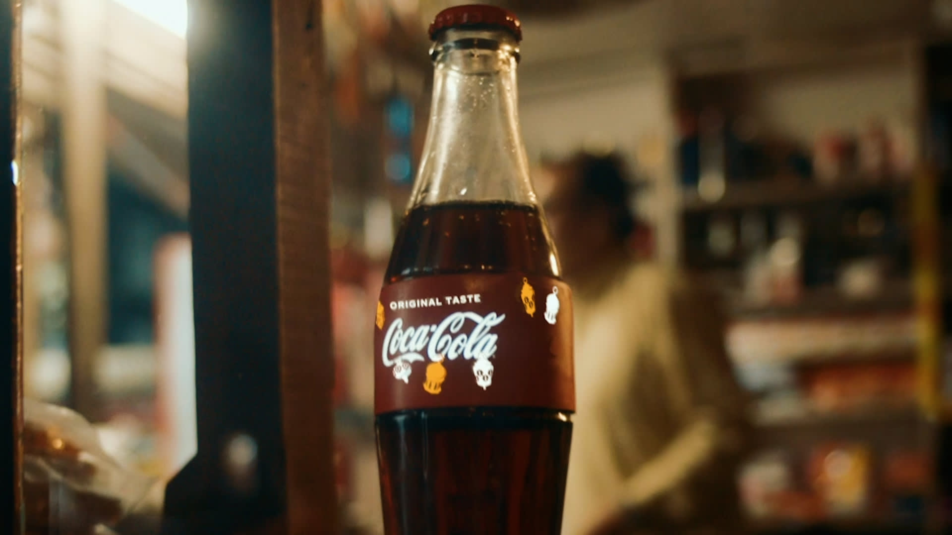 A classic glass Coca-Cola bottle with a red cap is placed on a counter in a dimly lit store, with shelves and a blurred figure in the background.