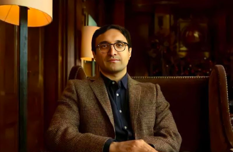 A man with glasses and dark hair sits in an armchair wearing a brown blazer over a dark shirt. Warm lighting and wood-paneled walls create a cozy, sophisticated atmosphere.