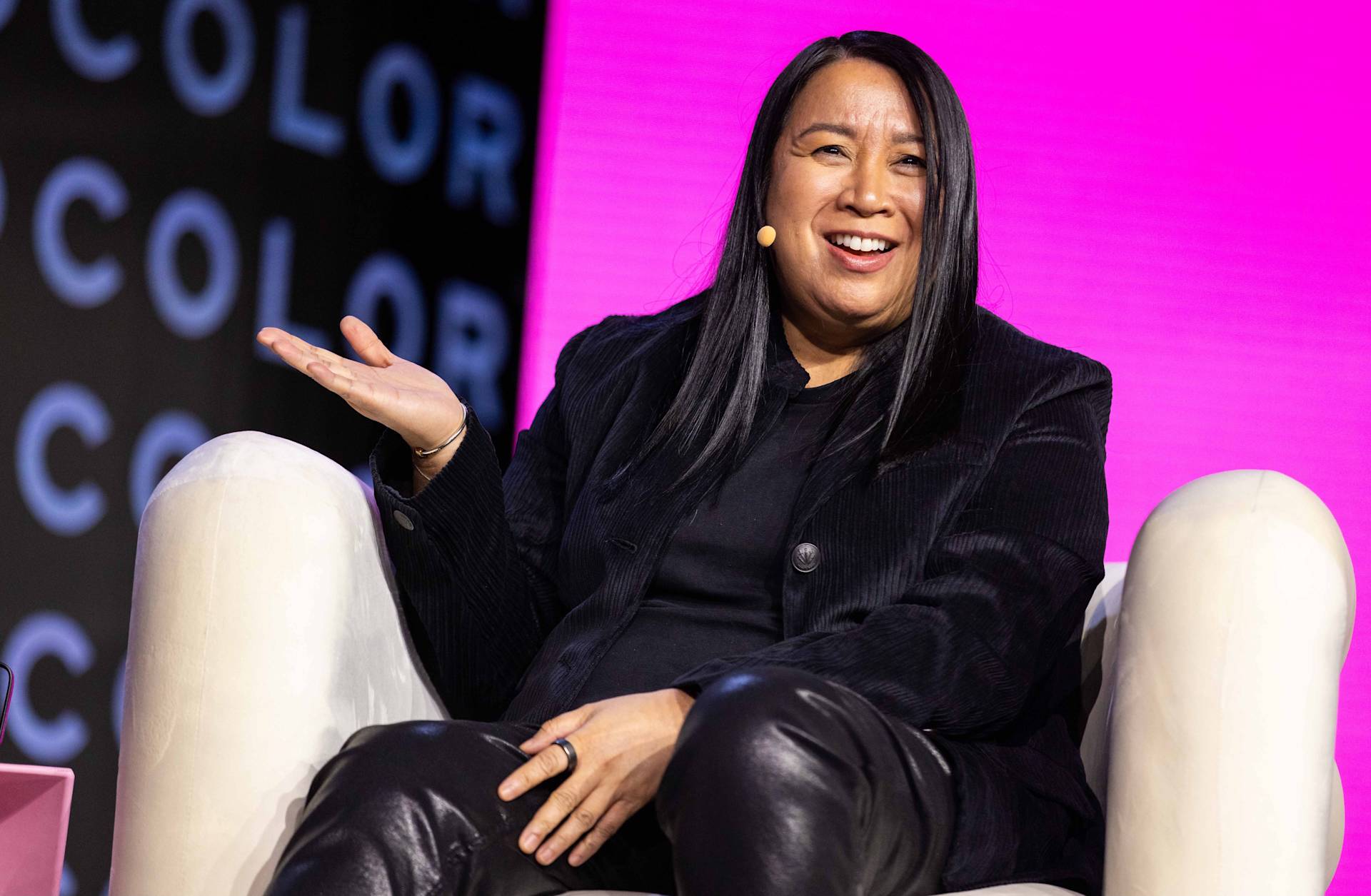 A person with long dark hair sits on a white chair, smiling and gesturing with one hand. They wear black clothing and have a microphone headset, with a vibrant pink and black background behind them.