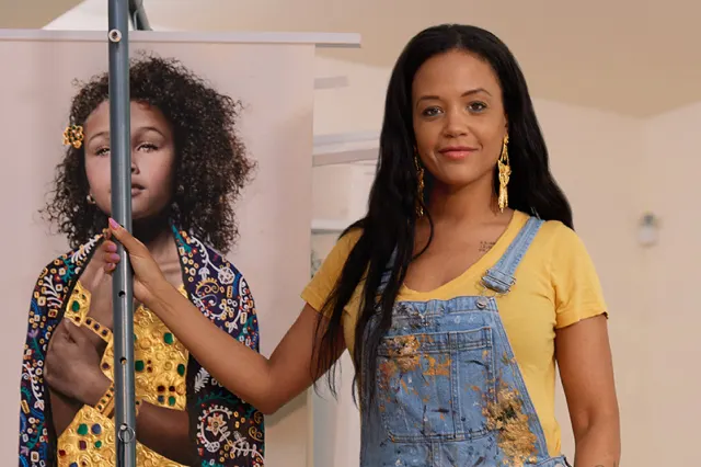 A woman in a yellow shirt and blue overalls with paint stains stands next to a portrait of a young girl with curly hair in a colorful robe. The woman is smiling and holding the edge of the portrait.