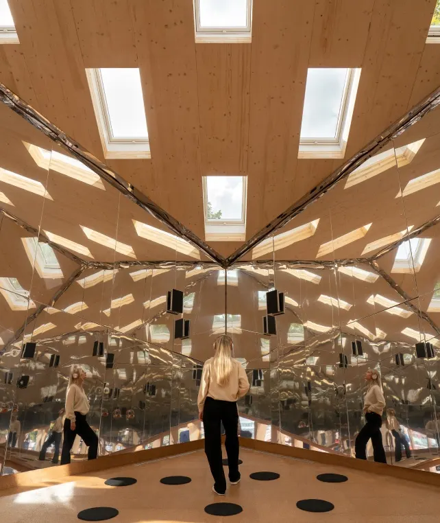 A person with long blond hair stands in a room with mirrored walls, reflecting their image multiple times. The ceiling is wooden with skylights, letting in natural light. Black dots are spaced on the floor.