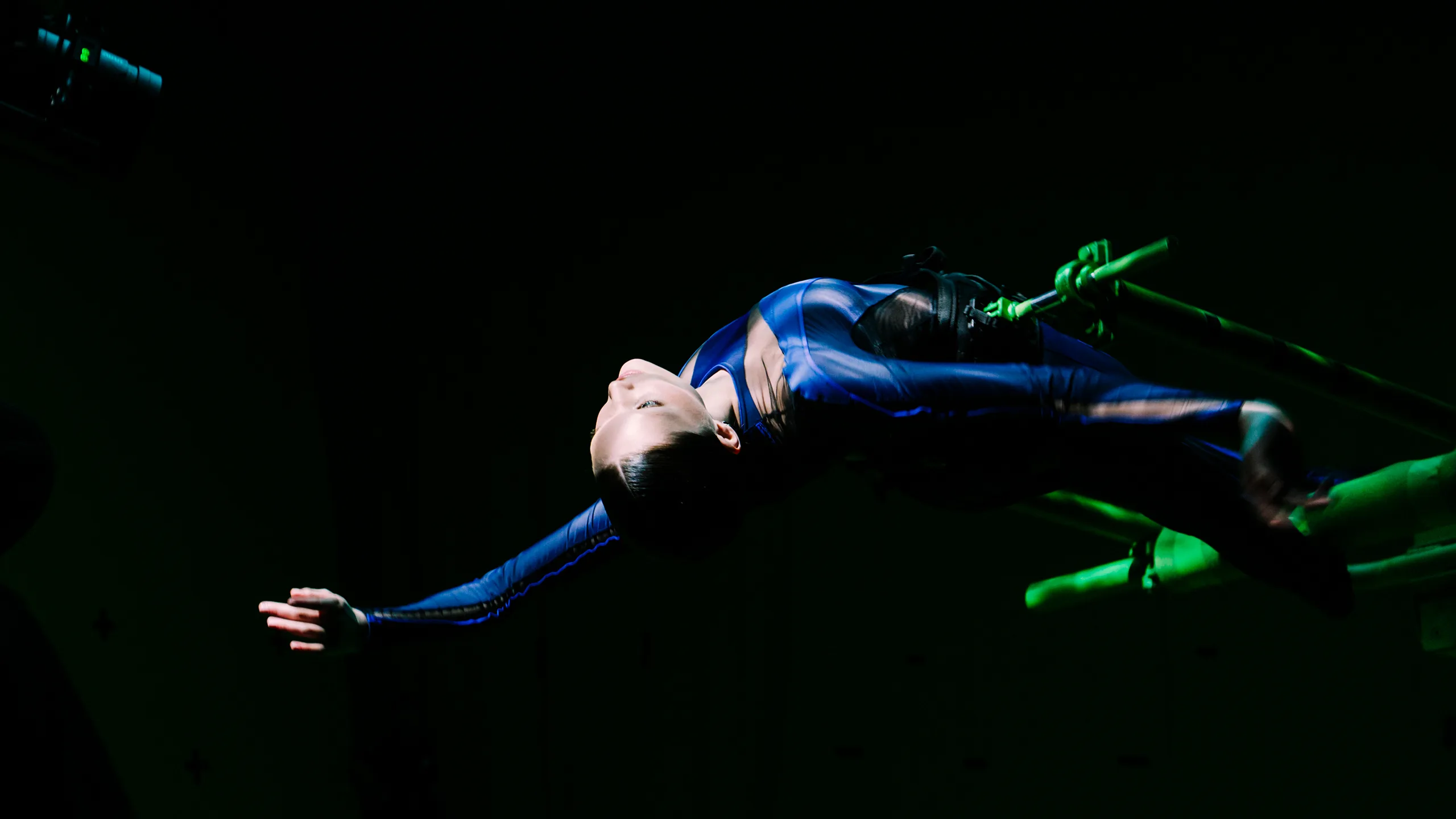 A person in a blue bodysuit performs an aerial acrobatic pose, arching their back and extending their arms while suspended from green apparatus against a dark background.