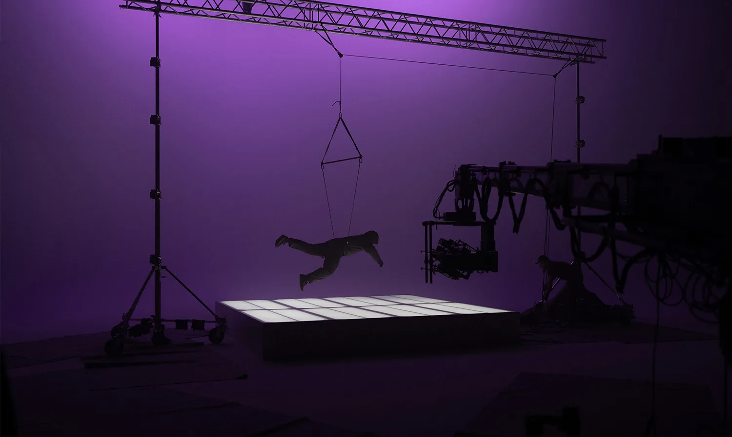 A person is suspended horizontally by a harness above a glowing, tiled platform in a studio with purple lighting. Film equipment and silhouettes of crew members are visible in the background.