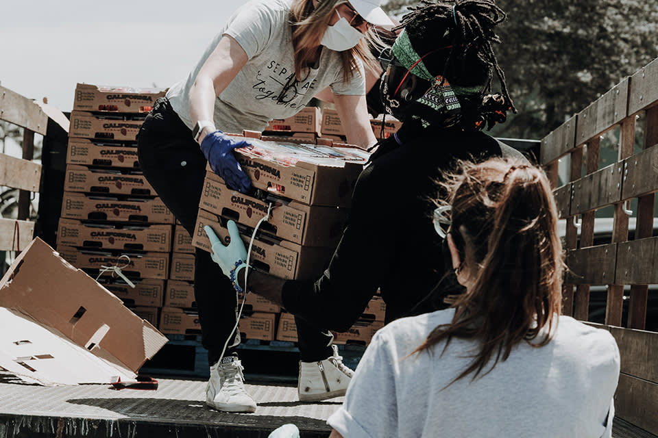 Three people wearing masks and gloves unload and pass boxes from a truck. One person hands a box to another while a third person sits nearby. Stacks of boxes are visible in the background. It appears to be a volunteer activity.