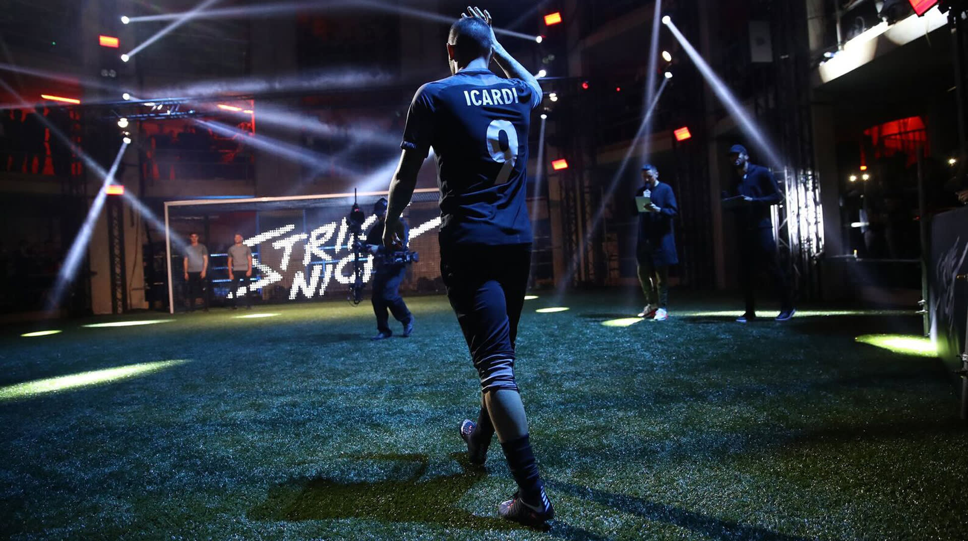A soccer player in a black jersey labeled ICARDI 9 walks on an indoor field toward a lit goal, with other players and a bright TRIAL NIGHT sign in the background.