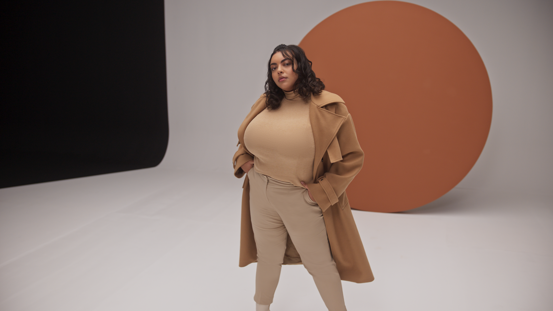 A confident woman in beige high-waisted pants, a matching turtleneck, and a long camel coat poses with hands in pockets in a minimalist studio with a large brown circle backdrop.