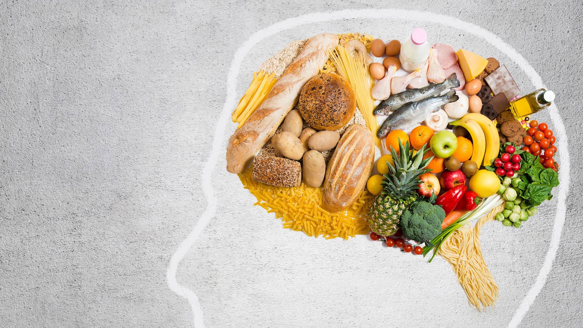 Various foods including bread, pasta, fish, eggs, fruits, vegetables, and oil are arranged inside an outline of a human head drawn on a gray surface, symbolizing the importance of nutrition for brain health.