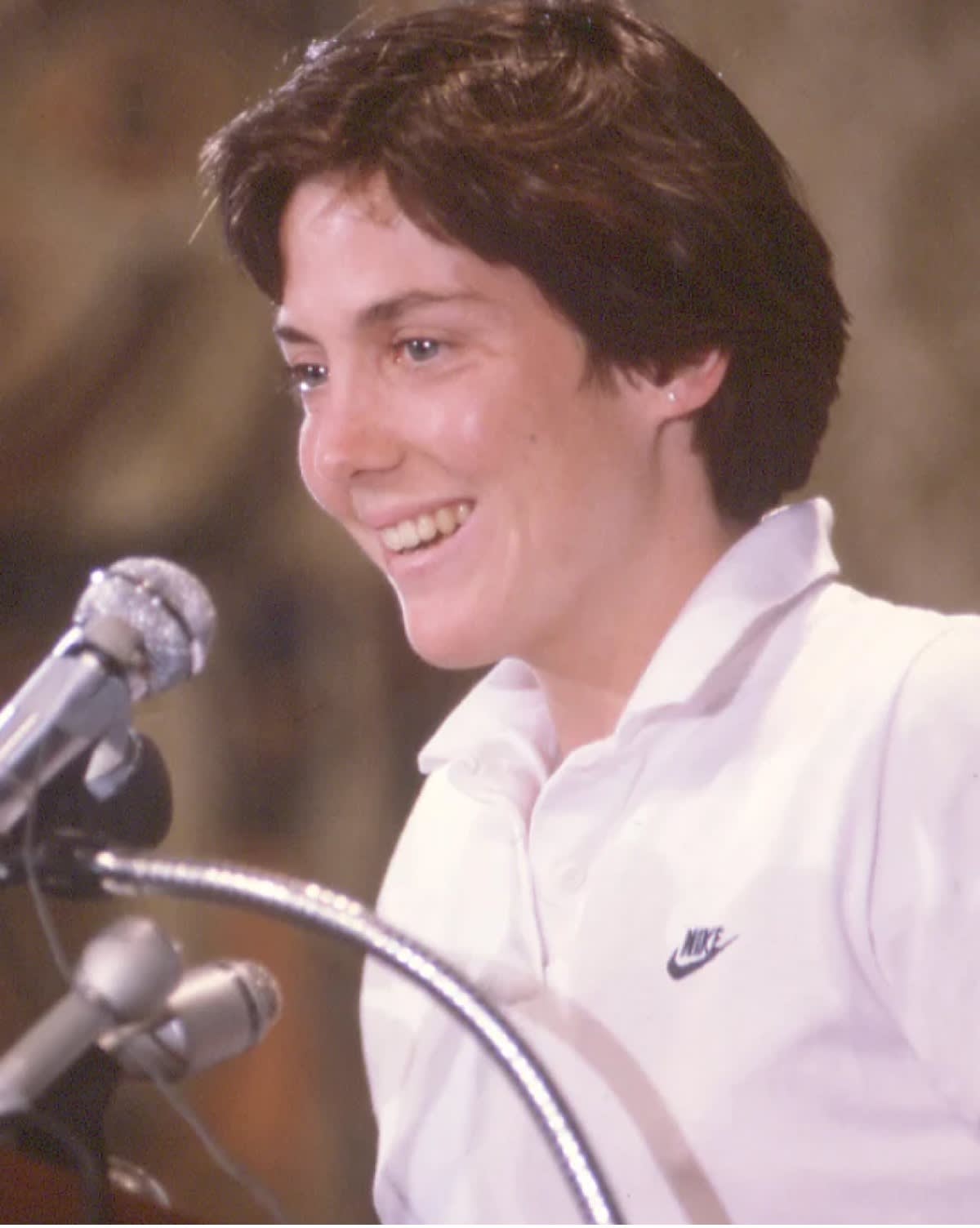 A person with short dark hair smiles while speaking at a microphone. They are wearing a white Nike polo shirt. The background is softly blurred.