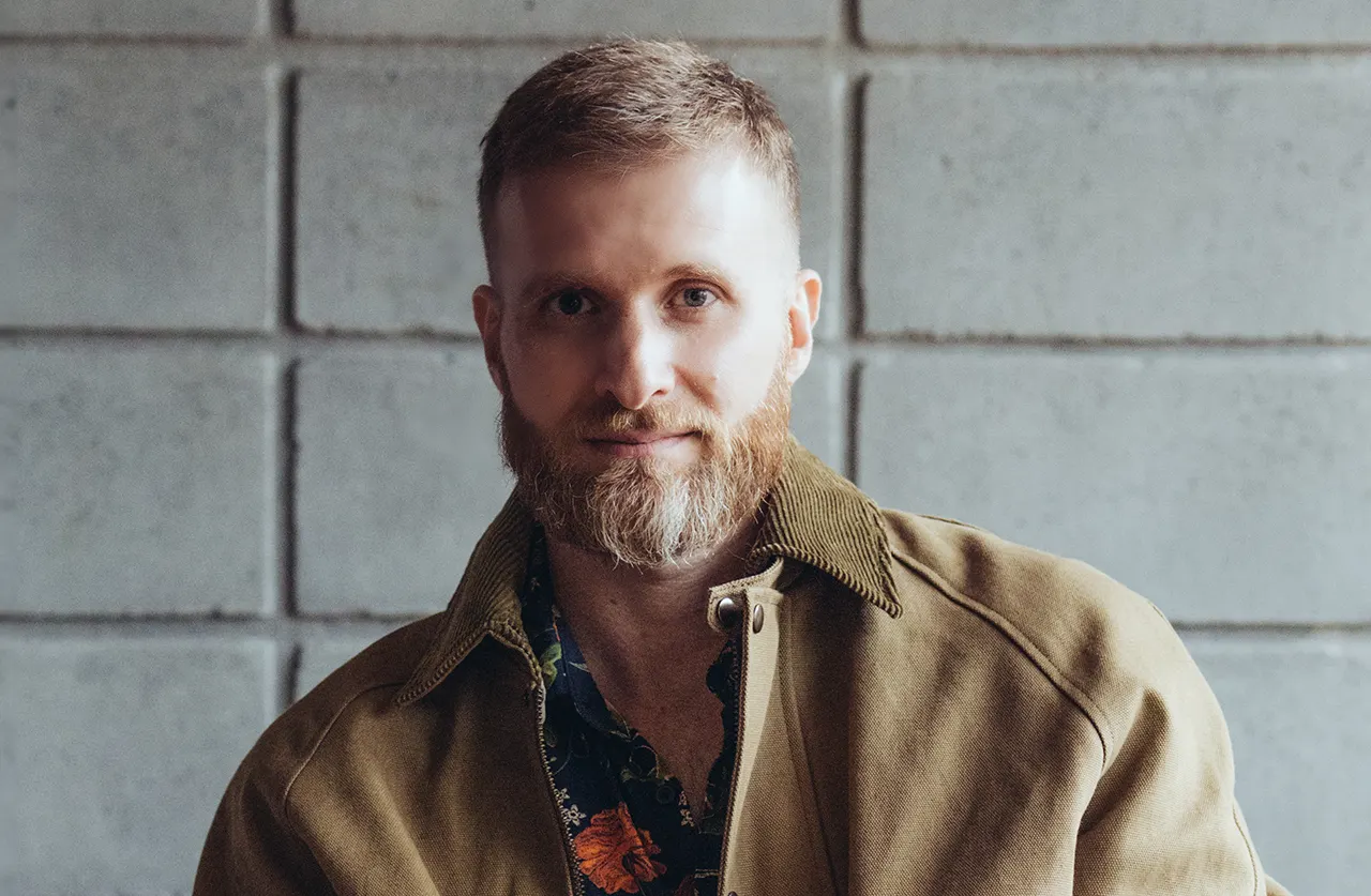 A man with short light brown hair and a full beard wears a tan jacket over a dark floral shirt, standing in front of a gray concrete block wall. He looks directly at the camera with a neutral expression.
