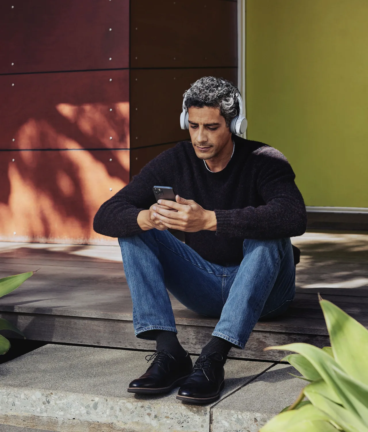 A man with curly hair sits on concrete steps, wearing headphones, a dark sweater, and jeans. He looks at his smartphone. Green plants and modern building panels are in the background.