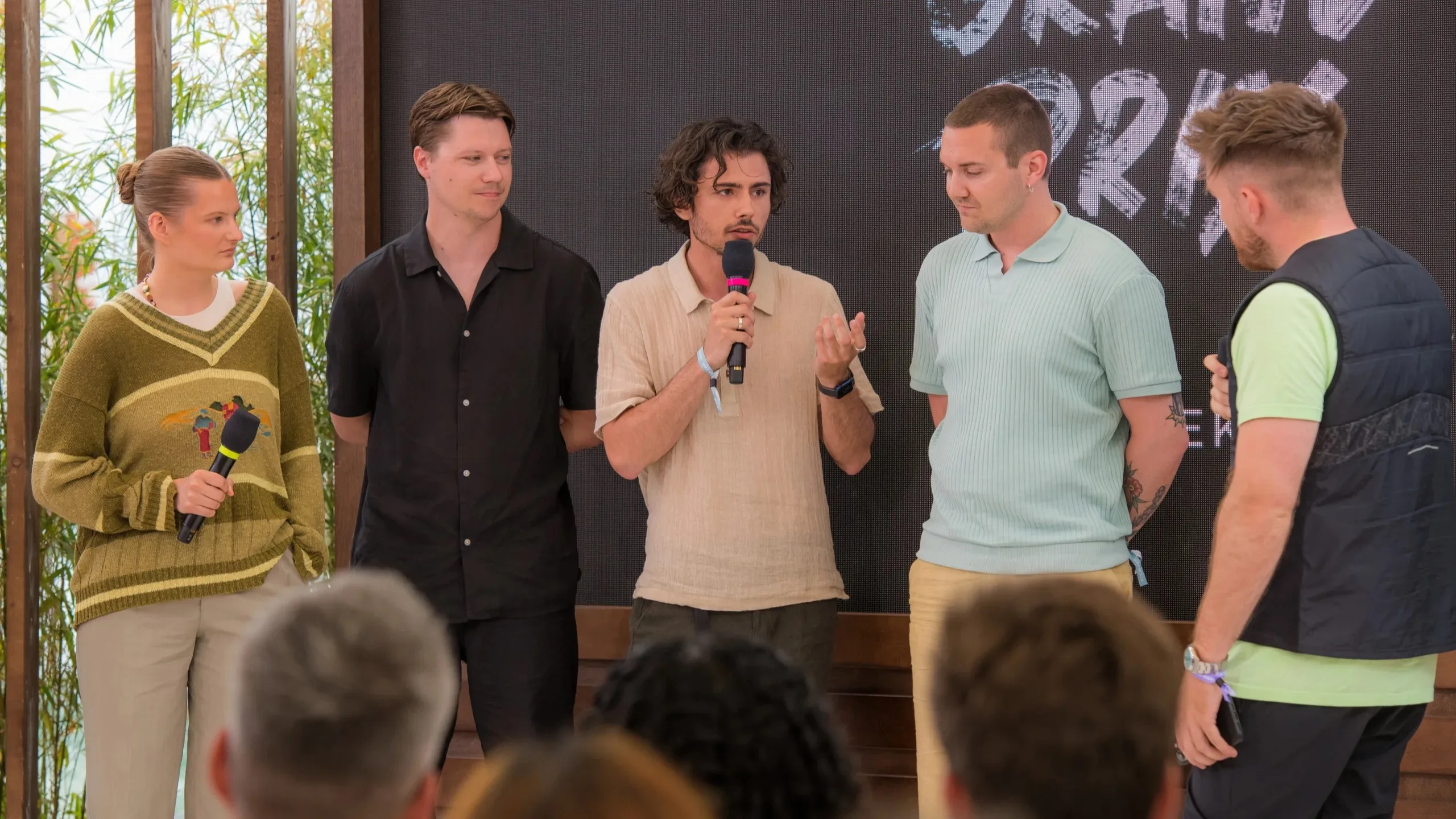 Five people stand in front of an audience. One person in the center speaks into a microphone, while the others listen. Theres a large screen behind them with partial text visible.
