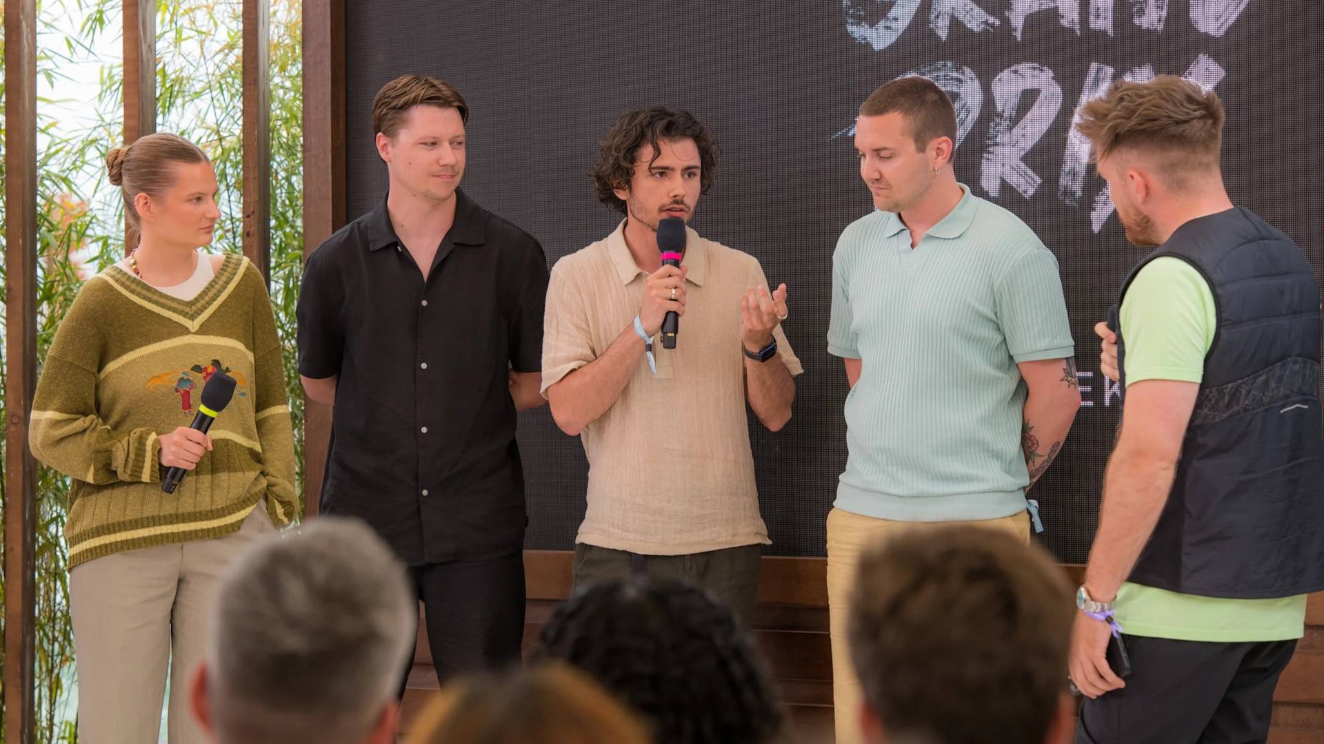 Five people stand in front of an audience. One person in the center speaks into a microphone, while the others listen. Theres a large screen behind them with partial text visible.
