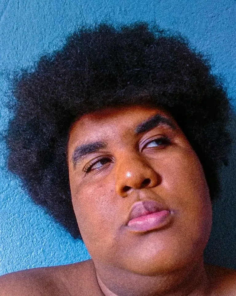 Person with short, curly black hair and medium brown skin looking off to the side with a neutral expression, against a textured blue wall background.