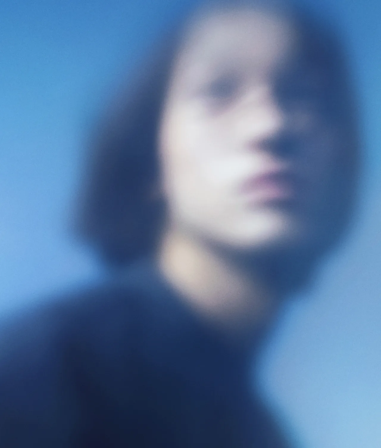 A blurred portrait of a person with short hair, wearing a dark top, set against a soft blue background. The face is out of focus, giving the image an abstract and dreamy appearance.
