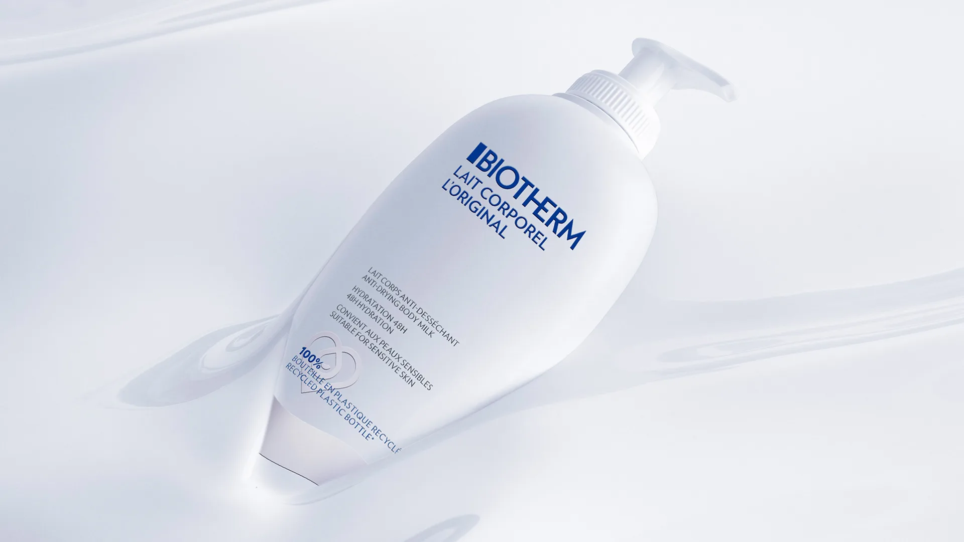 A white bottle of Biotherm Lait Corporel L’Original body milk with a pump dispenser lies on a light, glossy surface. The bottle features blue and gray text and a subtle wave graphic.
