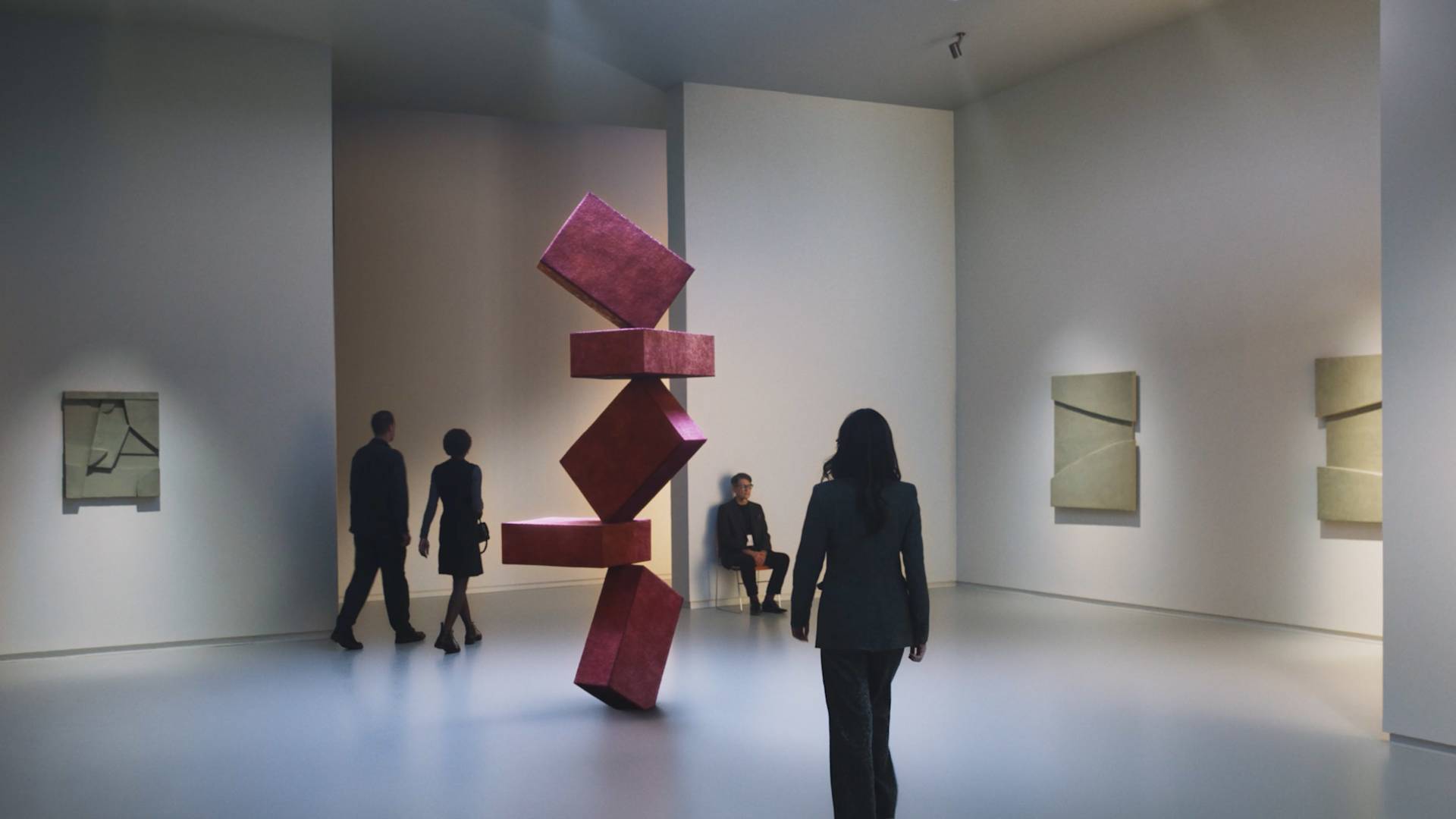 People view abstract art in a modern gallery, featuring a large red geometric sculpture in the center and minimalist artworks on the walls, with soft lighting and a spacious, white interior.