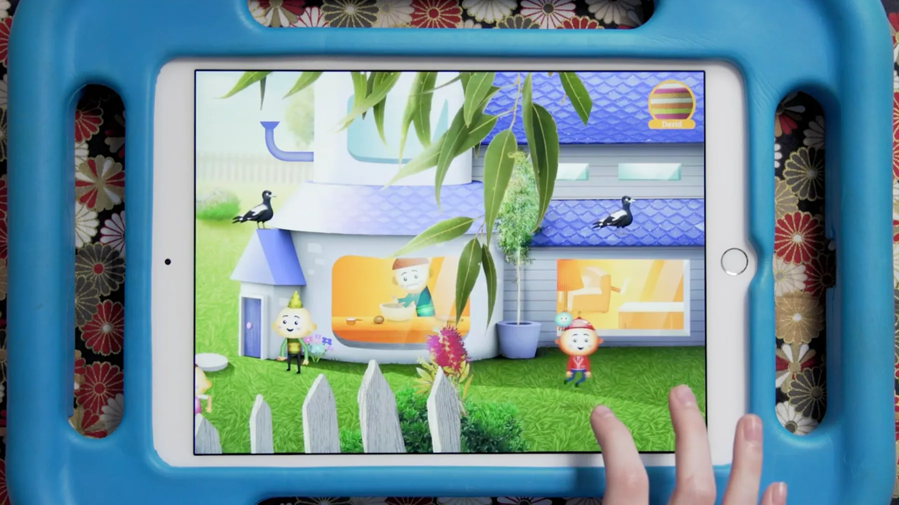 A childs hand holds a blue tablet displaying a colorful animated house scene with cartoon characters outside, birds on the roof, and green grass, surrounded by a floral-patterned background.