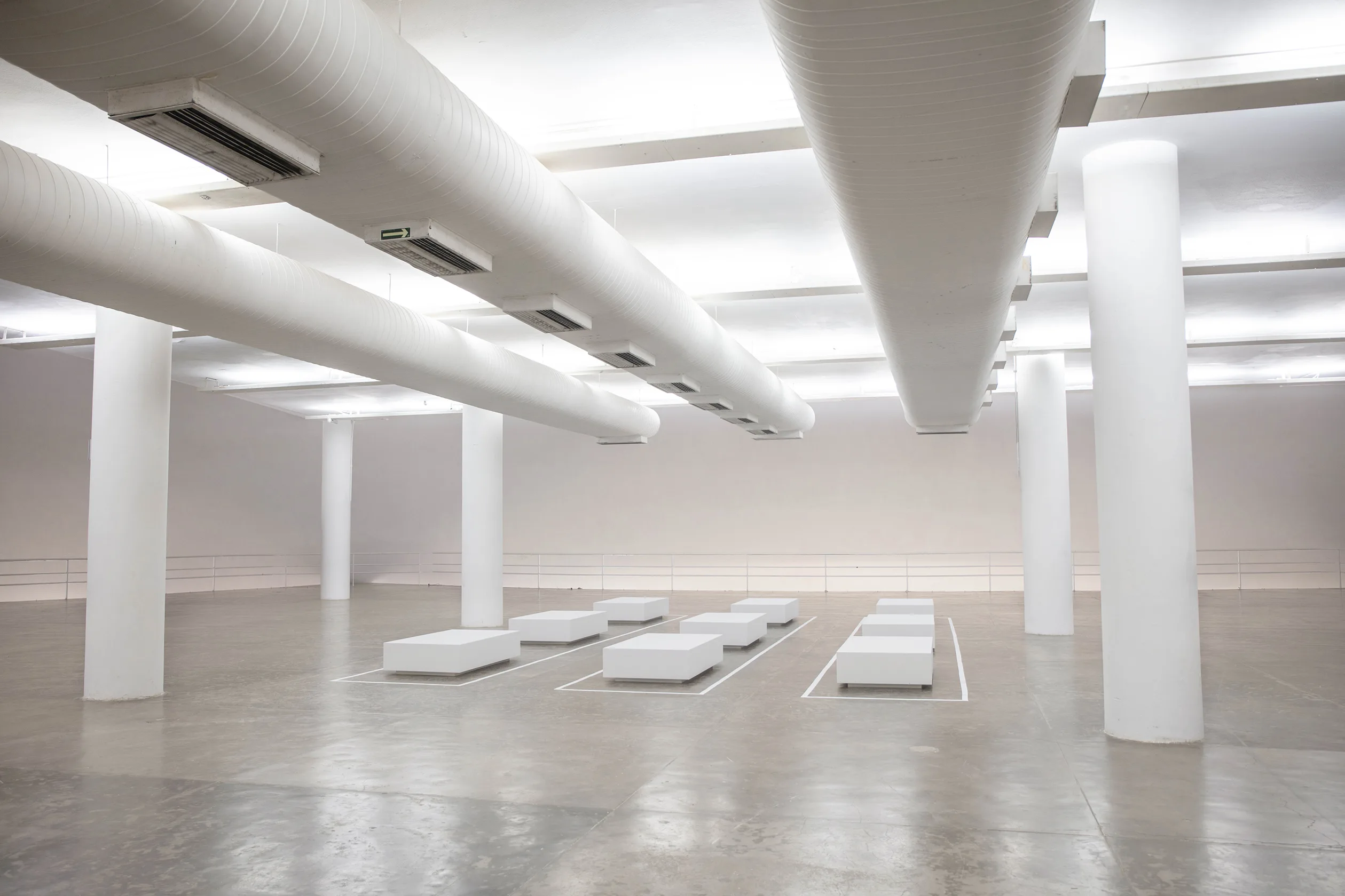 A spacious, minimalist room with large white columns, exposed ventilation ducts on the ceiling, and rows of low rectangular white platforms arranged on a polished concrete floor.