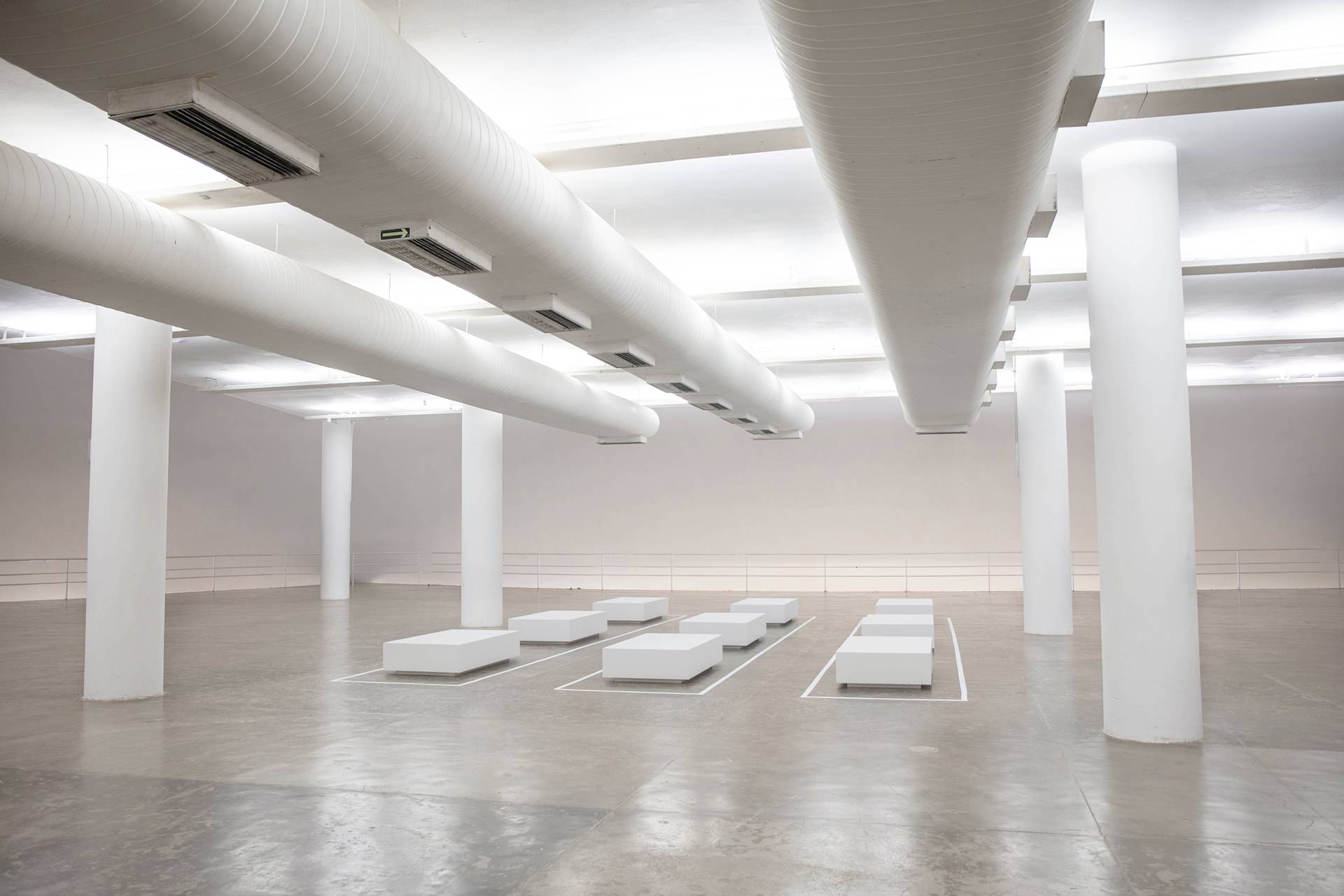 A spacious, minimalist room with large white columns, exposed ventilation ducts on the ceiling, and rows of low rectangular white platforms arranged on a polished concrete floor.