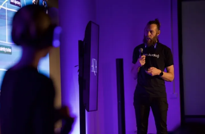 A man with a beard, wearing headphones and holding a microphone, speaks in front of a screen during a presentation. Another person in the foreground listens. The room is lit with purple lighting.