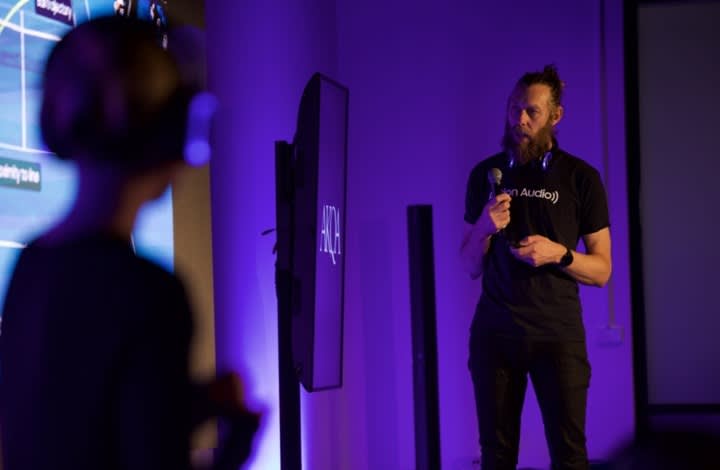 A man with a beard, wearing headphones and holding a microphone, speaks in front of a screen during a presentation. Another person in the foreground listens. The room is lit with purple lighting.