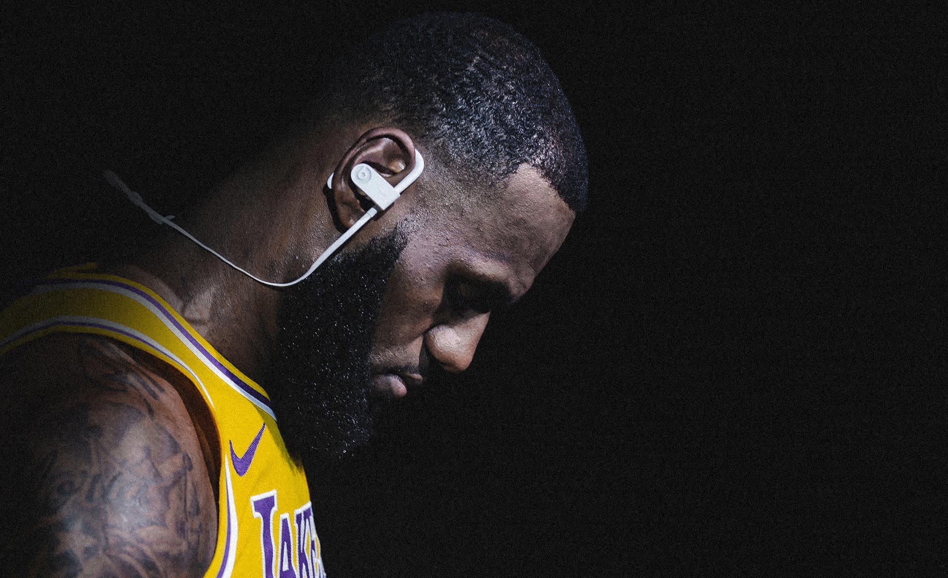 A basketball player in a yellow Lakers jersey looks down in deep concentration, wearing wireless headphones against a dark background with dramatic lighting.