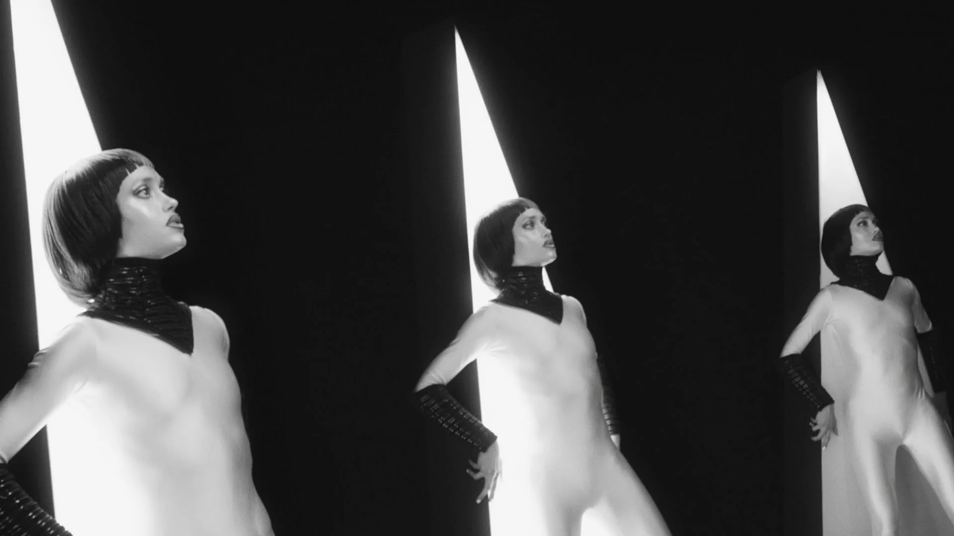 Three women in futuristic, form-fitting outfits stand in front of vertical beams of light in a black-and-white setting. Each has short hair and a high-necked collar, striking confident poses against a dark background.