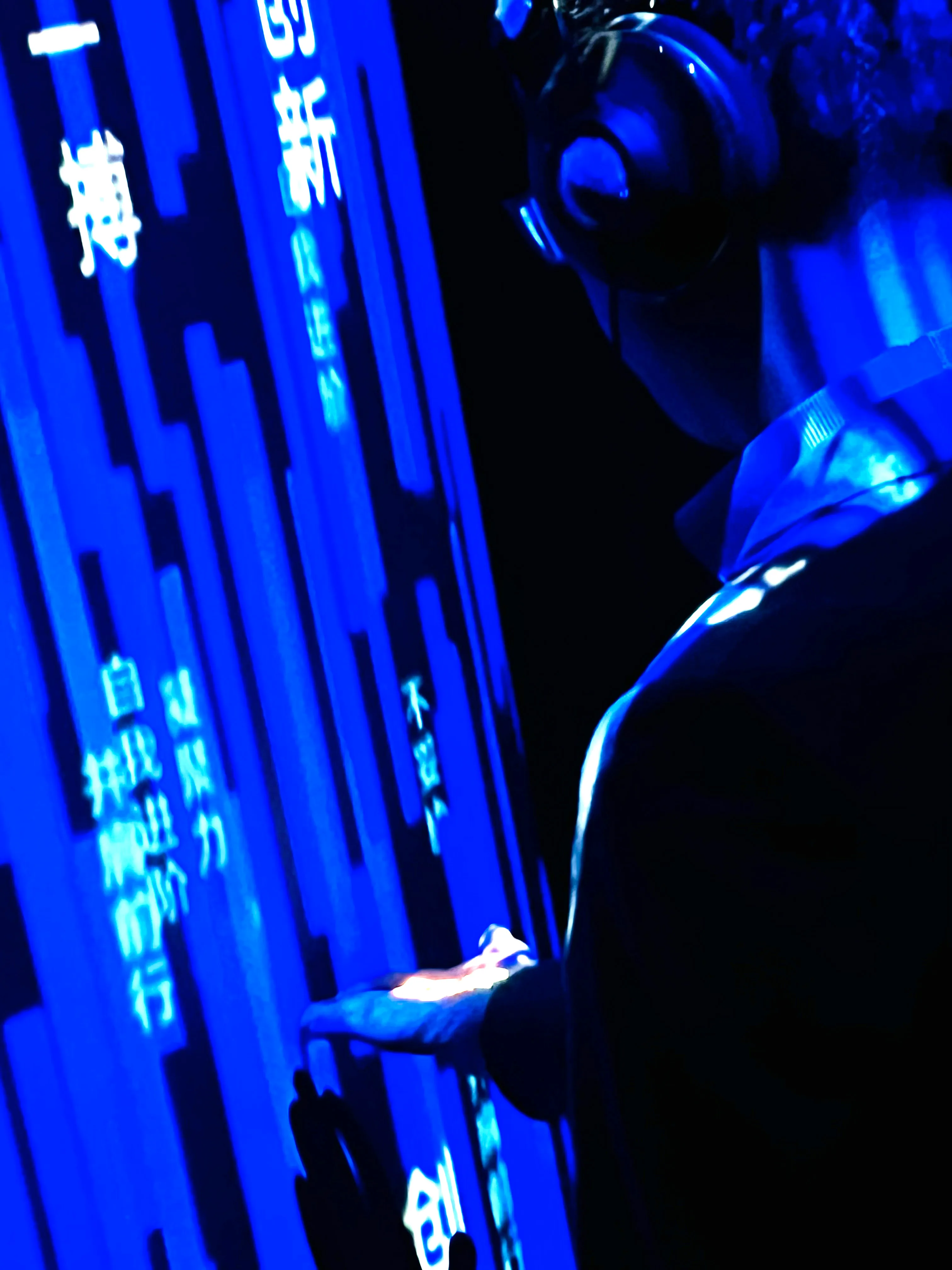 A person wearing headphones interacts with a digital touchscreen displaying blue-lit vertical lines and Chinese characters in a dark environment. Their hand and part of their face are illuminated by the screen’s glow.