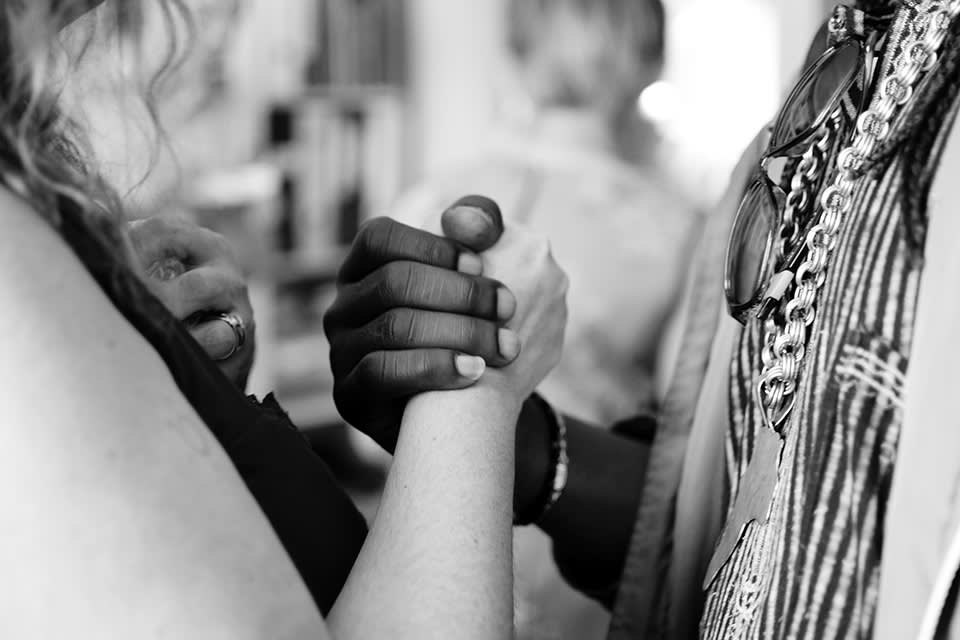 Two people of different skin tones clasp hands in a firm handshake, emphasizing unity and connection. The image is black and white, with both individuals partially visible, and blurred background elements.