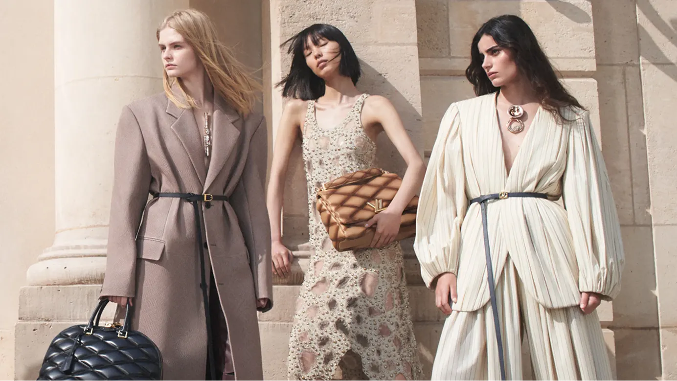 Three models stand in front of stone columns wearing designer clothing and carrying quilted handbags.