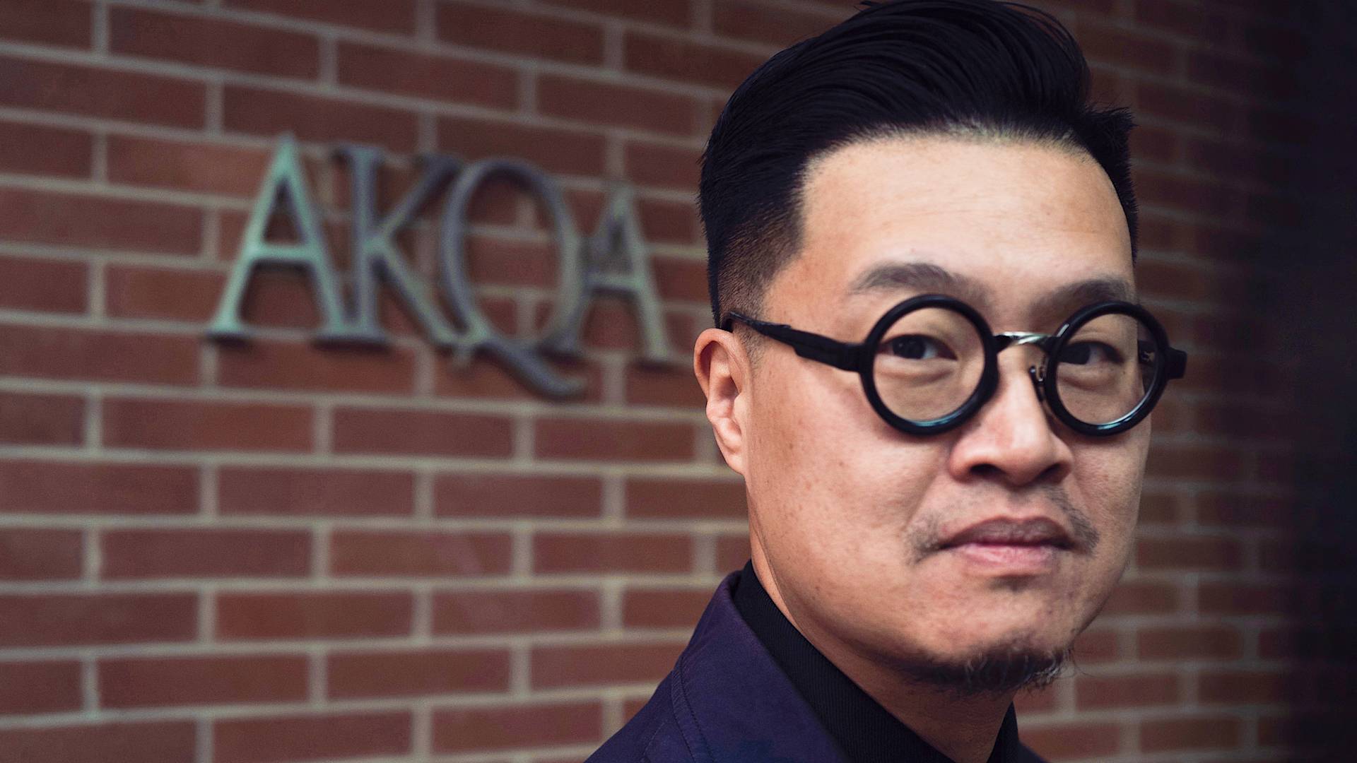 A person with short dark hair, round black glasses, and a trimmed beard stands in front of a brick wall with the letters AKQA displayed on it.